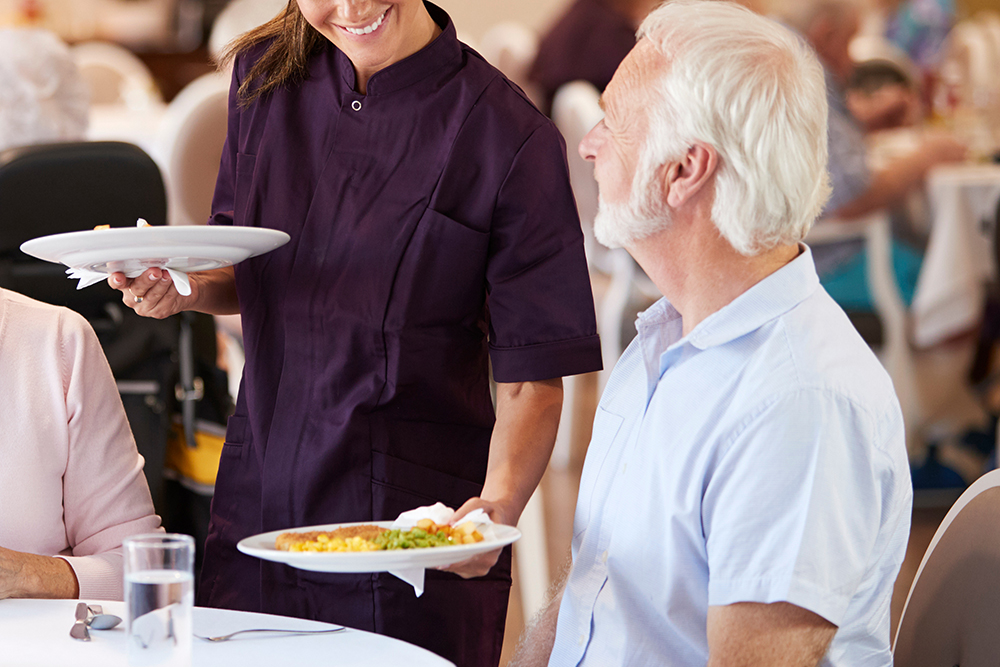 Senior Couple Being Served With Meal By Carer In Dining Room Of Retirement Home